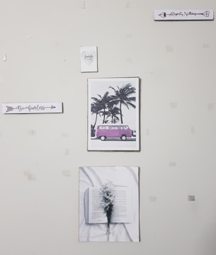 A wall with five pieces of art decor including a sign that says 'Be fearless', a picture of a pink van by palm trees, a sign that says 'Regret Nothing', a picture of an open book with flowers, and a small sign that says 'family forever'. The wall appears to have some tape or marks.
