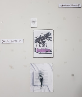 A wall with five pieces of art decor including a sign that says 'Be fearless', a picture of a pink van by palm trees, a sign that says 'Regret Nothing', a picture of an open book with flowers, and a small sign that says 'family forever'. The wall appears to have some tape or marks.