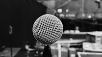 A close-up of a microphone with a metal mesh grille appears prominently in the foreground, with a blurred background depicting what seems to be a stage or backstage area. The image is in black and white, emphasizing the texture of the microphone and creating a moody atmosphere.