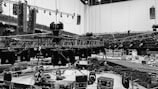 An indoor scene featuring a large, organized setup of stage rigging and equipment during an apparent stage construction or preparation. Metal trusses are suspended from the ceiling with numerous lighting fixtures attached. Several large speakers are visible and various boxes and cases are scattered on the floor, indicating a technical setup for an event.