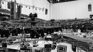 An indoor scene featuring a large, organized setup of stage rigging and equipment during an apparent stage construction or preparation. Metal trusses are suspended from the ceiling with numerous lighting fixtures attached. Several large speakers are visible and various boxes and cases are scattered on the floor, indicating a technical setup for an event.