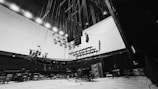 A large, empty stage setup with numerous lights and speakers hanging from the ceiling. The stage equipment is positioned in front of a high wall, with various cables and rigging tools visible. Black curtains and several equipment cases are arranged around the floor, giving the space an industrial feel.