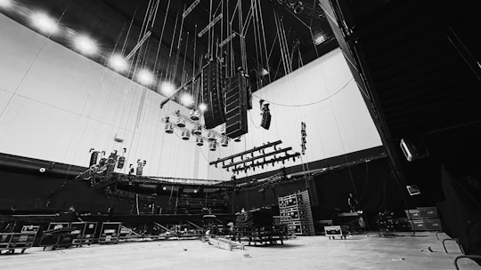 A large, empty stage setup with numerous lights and speakers hanging from the ceiling. The stage equipment is positioned in front of a high wall, with various cables and rigging tools visible. Black curtains and several equipment cases are arranged around the floor, giving the space an industrial feel.