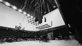 A large, empty stage setup with numerous lights and speakers hanging from the ceiling. The stage equipment is positioned in front of a high wall, with various cables and rigging tools visible. Black curtains and several equipment cases are arranged around the floor, giving the space an industrial feel.