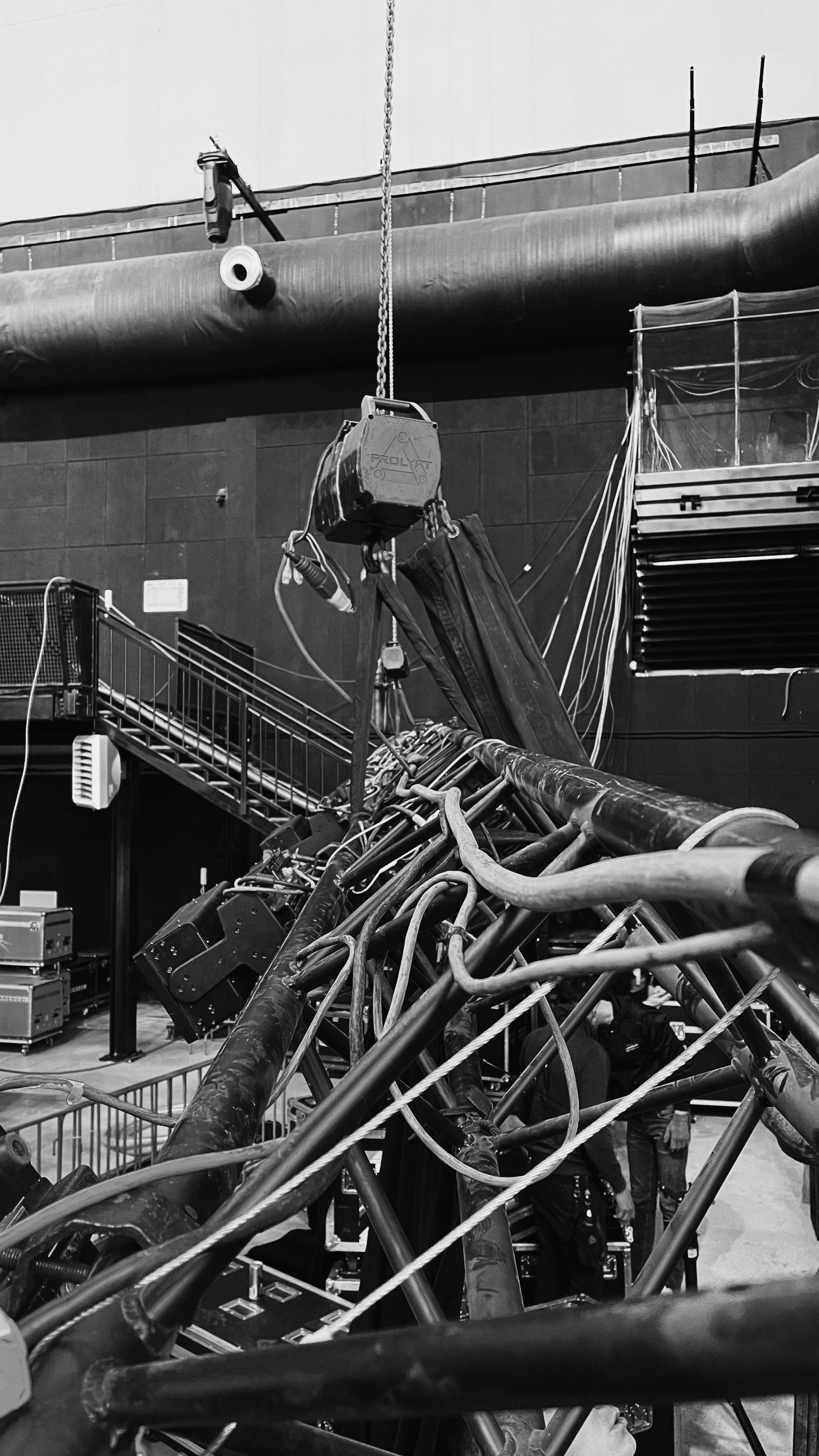 An industrial indoor scene showing an intricate setup of machinery and equipment. There are numerous cables and pipes intertwined, with a chain hoist hanging from above. A staircase is visible on the left side, leading to a platform with railings. Various electrical equipment and control boxes are positioned around the area. The environment seems to be part of a larger facility, with a large vent pipe running horizontally across the wall in the background.