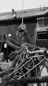 An industrial indoor scene showing an intricate setup of machinery and equipment. There are numerous cables and pipes intertwined, with a chain hoist hanging from above. A staircase is visible on the left side, leading to a platform with railings. Various electrical equipment and control boxes are positioned around the area. The environment seems to be part of a larger facility, with a large vent pipe running horizontally across the wall in the background.