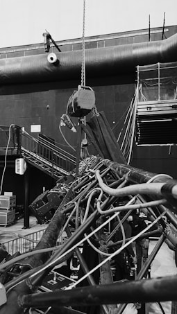 An industrial indoor scene showing an intricate setup of machinery and equipment. There are numerous cables and pipes intertwined, with a chain hoist hanging from above. A staircase is visible on the left side, leading to a platform with railings. Various electrical equipment and control boxes are positioned around the area. The environment seems to be part of a larger facility, with a large vent pipe running horizontally across the wall in the background.