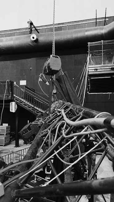An industrial indoor scene showing an intricate setup of machinery and equipment. There are numerous cables and pipes intertwined, with a chain hoist hanging from above. A staircase is visible on the left side, leading to a platform with railings. Various electrical equipment and control boxes are positioned around the area. The environment seems to be part of a larger facility, with a large vent pipe running horizontally across the wall in the background.