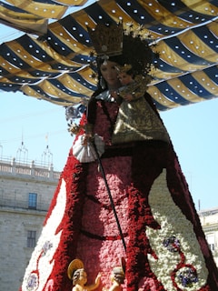 A large statue of a crowned figure holding a child is draped in a floral cloak, made of red, pink, and white flowers. The scene is set under a decorative canopy with blue and yellow striped fabric. In the foreground, smaller figures resembling angels or cherubs are visible.