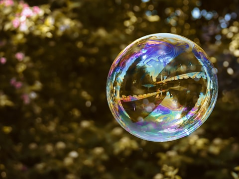 A large, iridescent soap bubble is floating in the air against a blurred background of greenery. The bubble reflects various colors and patterns, including some landscape-like reflections.