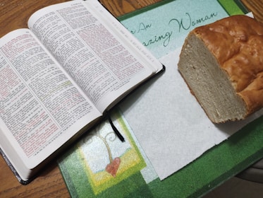 An open Bible with highlighted text in red and black lays on a table next to a loaf of bread on a paper towel. The table is decorated with a mat featuring a heart design and the phrase 'Amazing Woman.'