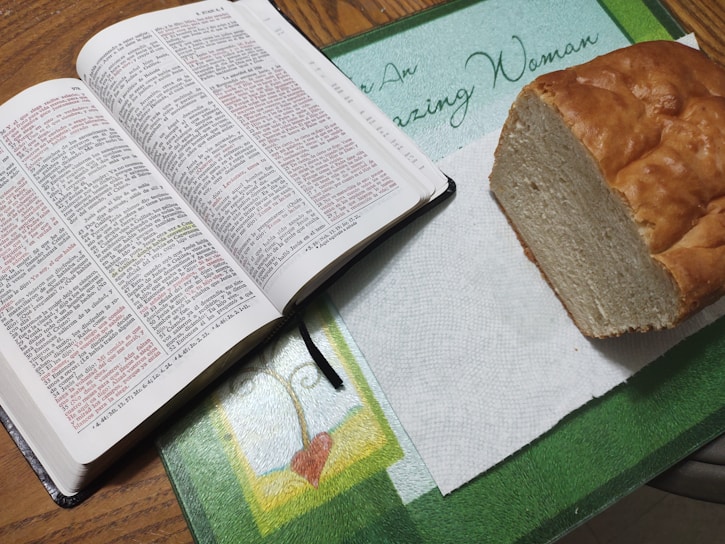 An open Bible with highlighted text in red and black lays on a table next to a loaf of bread on a paper towel. The table is decorated with a mat featuring a heart design and the phrase 'Amazing Woman.'
