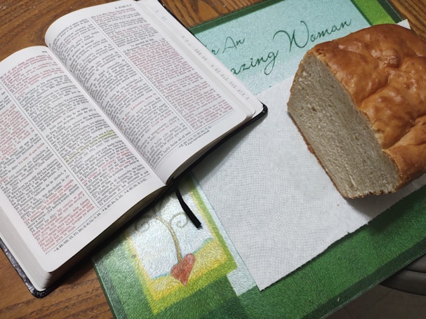 An open Bible with highlighted text in red and black lays on a table next to a loaf of bread on a paper towel. The table is decorated with a mat featuring a heart design and the phrase 'Amazing Woman.'