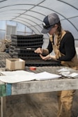 A person wearing overalls and a cap is working at a wooden table inside a greenhouse. They are focused on handling a seedling tray, using a small tool. The table is cluttered with various items, including papers, a notebook, a roll of tape, and a box. Stacks of soil trays are also visible in the background.