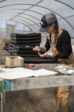 A person wearing overalls and a cap is working at a wooden table inside a greenhouse. They are focused on handling a seedling tray, using a small tool. The table is cluttered with various items, including papers, a notebook, a roll of tape, and a box. Stacks of soil trays are also visible in the background.