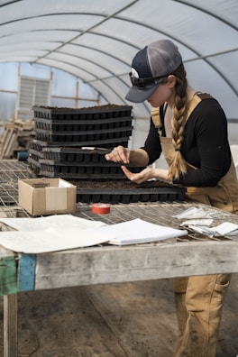A person wearing overalls and a cap is working at a wooden table inside a greenhouse. They are focused on handling a seedling tray, using a small tool. The table is cluttered with various items, including papers, a notebook, a roll of tape, and a box. Stacks of soil trays are also visible in the background.