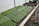 Trays of small green seedlings are organized neatly on a wooden table inside a greenhouse. A watering hose lies coiled next to the seedlings, and the environment appears to be set up for cultivating plants. The background includes additional trays, some filled with soil, indicating a plant nursery setup.