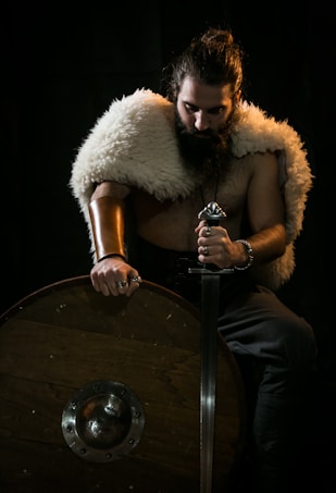 A person with long hair and a beard is sitting in a dimly lit setting, wearing a fur cloak over their shoulders and holding a sword. The individual rests one arm on a large round wooden shield featuring a metal boss. Rings adorn the person's fingers, adding to the warrior-like appearance.
