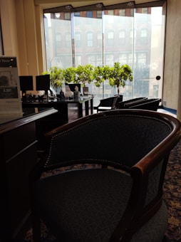 This is an indoor setting with several wooden chairs and a desk. There are plants placed on the desk, and a large window allows natural light to come into the room. The window shows a view of a brick building exterior. There is a sign on the desk displaying an interest rate, and two computer monitors are visible on the desk.