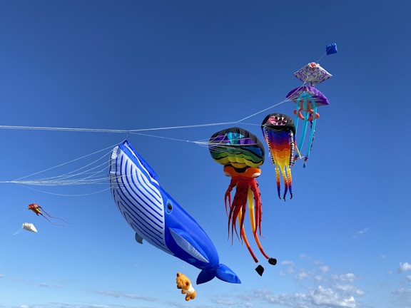 A collection of colorful kites, including a large blue whale, a rainbow-colored jellyfish, and a variety of other vibrant and uniquely shaped kites, soar against a clear blue sky.