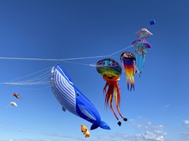 A collection of colorful kites, including a large blue whale, a rainbow-colored jellyfish, and a variety of other vibrant and uniquely shaped kites, soar against a clear blue sky.