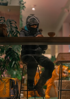 A person wearing a helmet and mask sits at a wooden counter in a cozy indoor setting. The individual appears to be looking at a device in their hands. In the background, there is lush foliage and a yellow delivery bag with the logo 'Glovo.' The lighting is warm and soft, enhancing the comfortable atmosphere.