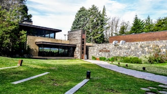 A modern, single-story building with large glass windows and a stone facade is surrounded by lush greenery and tall trees. A paved walkway leads to the building, and there is signage with the word 'Patagonia' on the stone wall. The overall setting includes a well-maintained lawn and various bushes and plants.