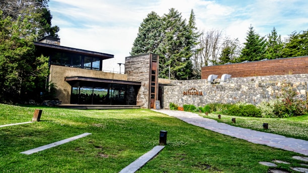 A modern, single-story building with large glass windows and a stone facade is surrounded by lush greenery and tall trees. A paved walkway leads to the building, and there is signage with the word 'Patagonia' on the stone wall. The overall setting includes a well-maintained lawn and various bushes and plants.
