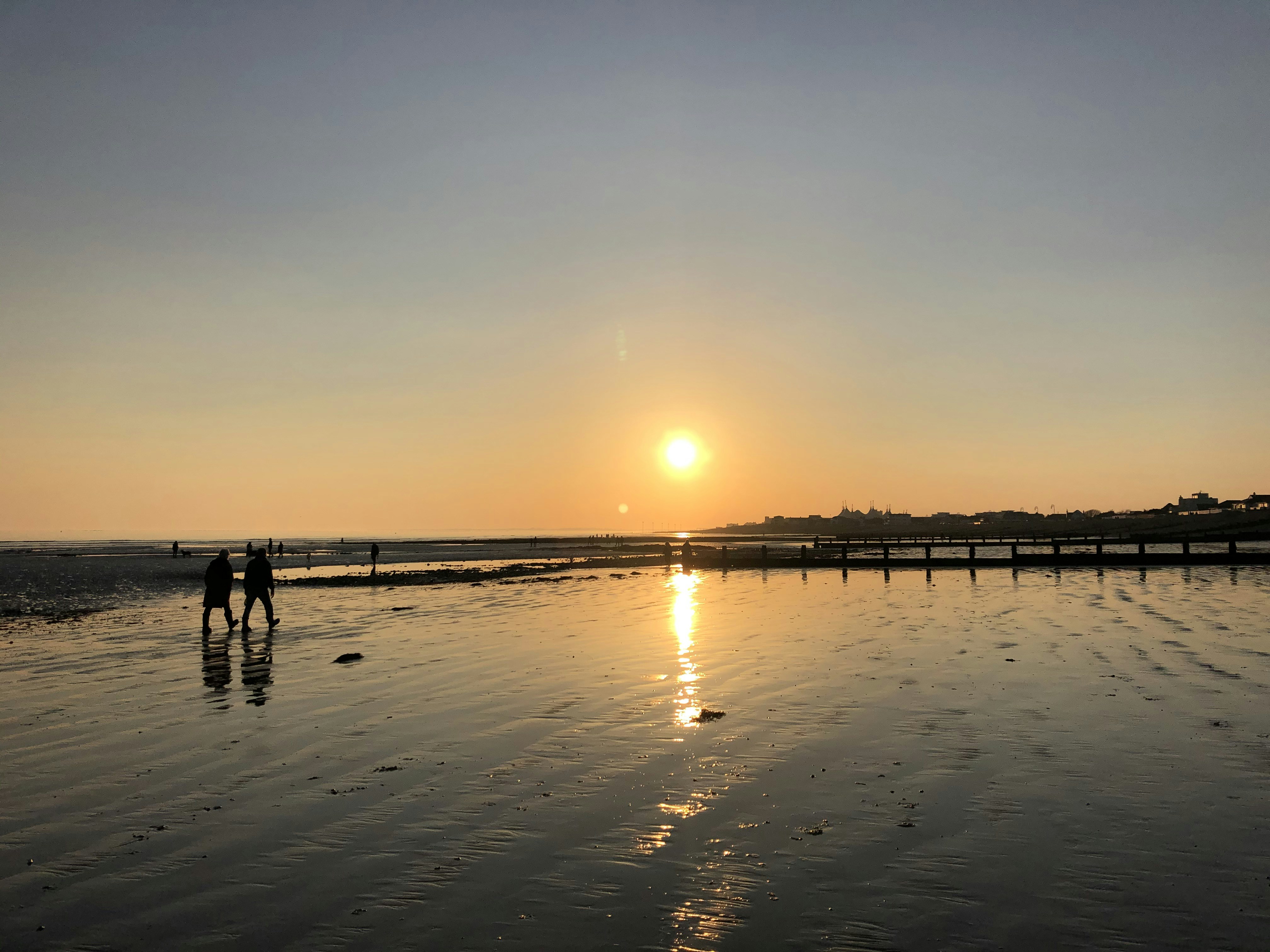 Couple walking on beach at sunset, golden reflections on wet sand, moody atmospheric sky, silhouette photography, dramatic lighting