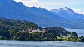 A large building with a red roof is nestled among lush green forests, with a serene lake in the foreground. Majestic mountains with snow-capped peaks rise in the background under a clear blue sky.