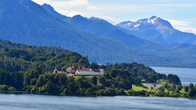 A large building with a red roof is nestled among lush green forests, with a serene lake in the foreground. Majestic mountains with snow-capped peaks rise in the background under a clear blue sky.