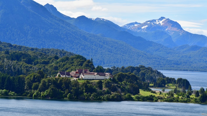A large building with a red roof is nestled among lush green forests, with a serene lake in the foreground. Majestic mountains with snow-capped peaks rise in the background under a clear blue sky.