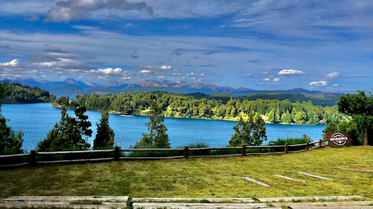 A scenic view of a lake surrounded by lush green forests and distant mountain ranges under a partly cloudy blue sky. There is a grassy foreground with a wooden fence and a sign that reads 'Patagonia' on the right side.