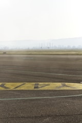 A foggy landscape with an airport runway in the foreground. The tarmac features painted yellow markings, and in the distance, industrial chimneys and faint outlines of trees can be seen.
