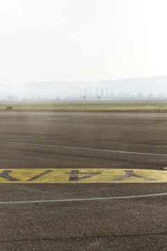 A foggy landscape with an airport runway in the foreground. The tarmac features painted yellow markings, and in the distance, industrial chimneys and faint outlines of trees can be seen.