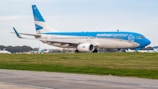 A large passenger airplane painted in blue and white with the logo of Aerolíneas Argentinas taxis on a runway. The background shows a clear sky and some distant trees, indicating an airport environment.