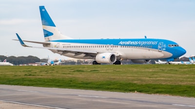 A large passenger airplane painted in blue and white with the logo of Aerolíneas Argentinas taxis on a runway. The background shows a clear sky and some distant trees, indicating an airport environment.
