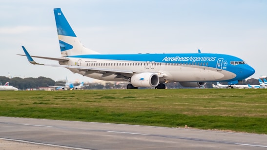 A large passenger airplane painted in blue and white with the logo of Aerolíneas Argentinas taxis on a runway. The background shows a clear sky and some distant trees, indicating an airport environment.