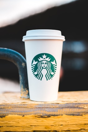 A white paper cup with a green logo is placed on a wooden surface with a blurred background. The cup features a recognizable brand symbol in its center.
