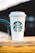 A white paper cup with a green logo is placed on a wooden surface with a blurred background. The cup features a recognizable brand symbol in its center.