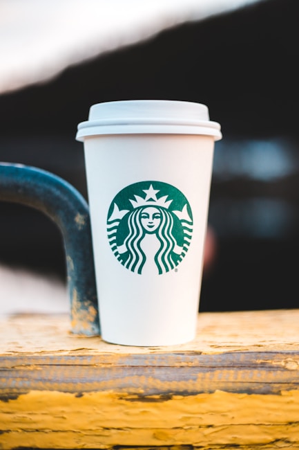 A white paper cup with a green logo is placed on a wooden surface with a blurred background. The cup features a recognizable brand symbol in its center.