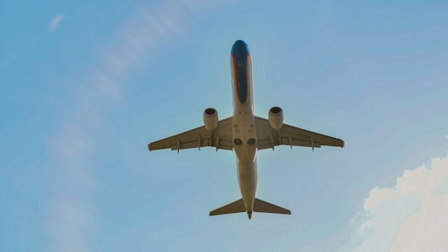 A commercial airplane is captured mid-flight from underneath, soaring through a clear blue sky. The aircraft appears sleek and modern, with visible jet engines and a distinct tail fin.