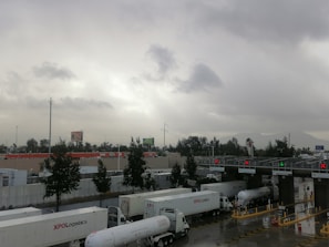 Several trucks are lined up at a toll station under a cloudy sky. The scene includes infrastructure like metal toll booths with lights, trees along the roadside, and advertising billboards in the background.