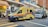 An ambulance with a distinctive yellow color and blue and red stripes is parked on a city street next to a small food stall. Several people are near the stall and a large commercial truck is parked behind the ambulance. The setting is an urban area with high-rise buildings and various signs, including a no-entry road sign and traffic lights.