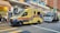 An ambulance with a distinctive yellow color and blue and red stripes is parked on a city street next to a small food stall. Several people are near the stall and a large commercial truck is parked behind the ambulance. The setting is an urban area with high-rise buildings and various signs, including a no-entry road sign and traffic lights.