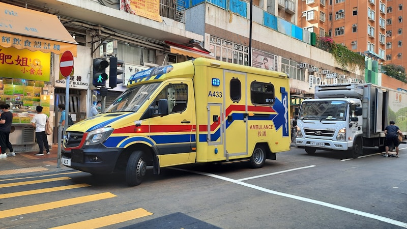 An ambulance with a distinctive yellow color and blue and red stripes is parked on a city street next to a small food stall. Several people are near the stall and a large commercial truck is parked behind the ambulance. The setting is an urban area with high-rise buildings and various signs, including a no-entry road sign and traffic lights.