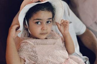 A young child with dark hair is wearing a delicate pink dress with floral embroidery and a white sun hat. The child is sitting on a person's lap, holding the brim of the hat with both hands, and looking directly at the camera with a calm expression.