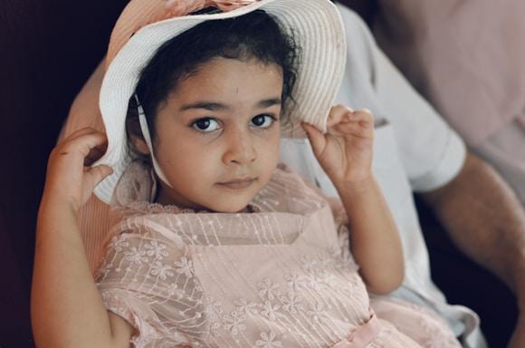 A young child with dark hair is wearing a delicate pink dress with floral embroidery and a white sun hat. The child is sitting on a person's lap, holding the brim of the hat with both hands, and looking directly at the camera with a calm expression.
