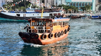 A wooden ferry boat is sailing on a body of water, with passengers visible inside. The boat is equipped with life buoys around the sides. In the background, larger boats are docked near a cityscape with tall buildings and greenery. Colorful flags are strung above, adding a festive atmosphere.