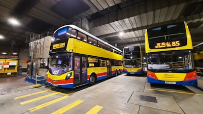 A parking area inside a bus depot houses several double-decker buses. The buses are brightly colored with yellow and blue shades and have digital destination displays, with one bus showing 'Haven Valley Rd 19' and another displaying 'Braemar Hill 85'. The depot has a concrete floor, high ceilings, and is illuminated with overhead lights. The area is organized with marked parking lines.